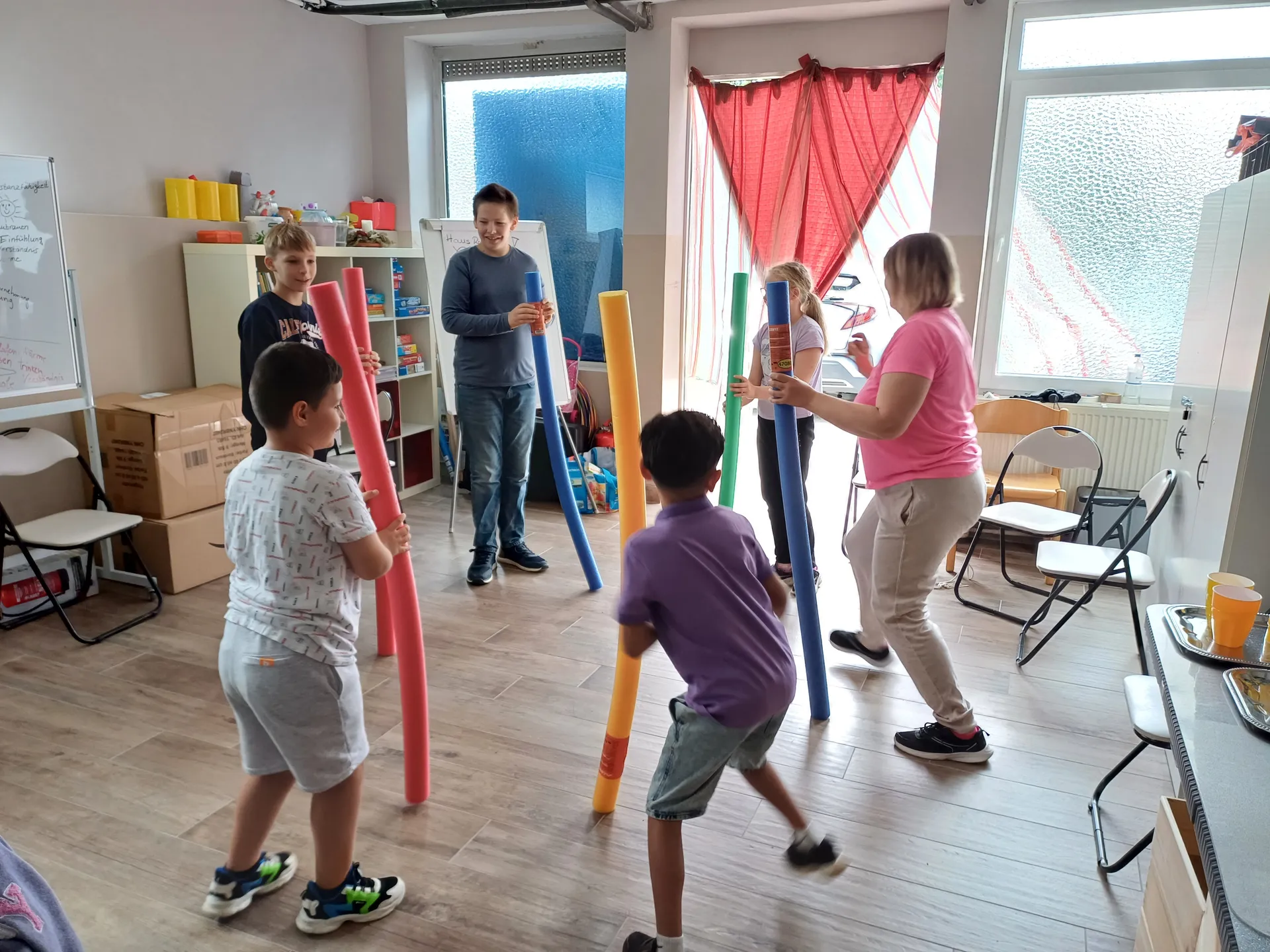 Kinder und eine Betreuerin bei einem Bewegungsspiel mit Schwimmnudeln im Gruppenraum
